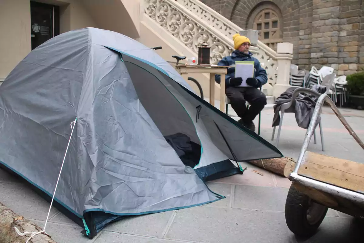 Una tienda de campaña está montada en una acera junto a una carretilla y una persona sentada en una silla con un gorro amarillo sosteniendo papeles.