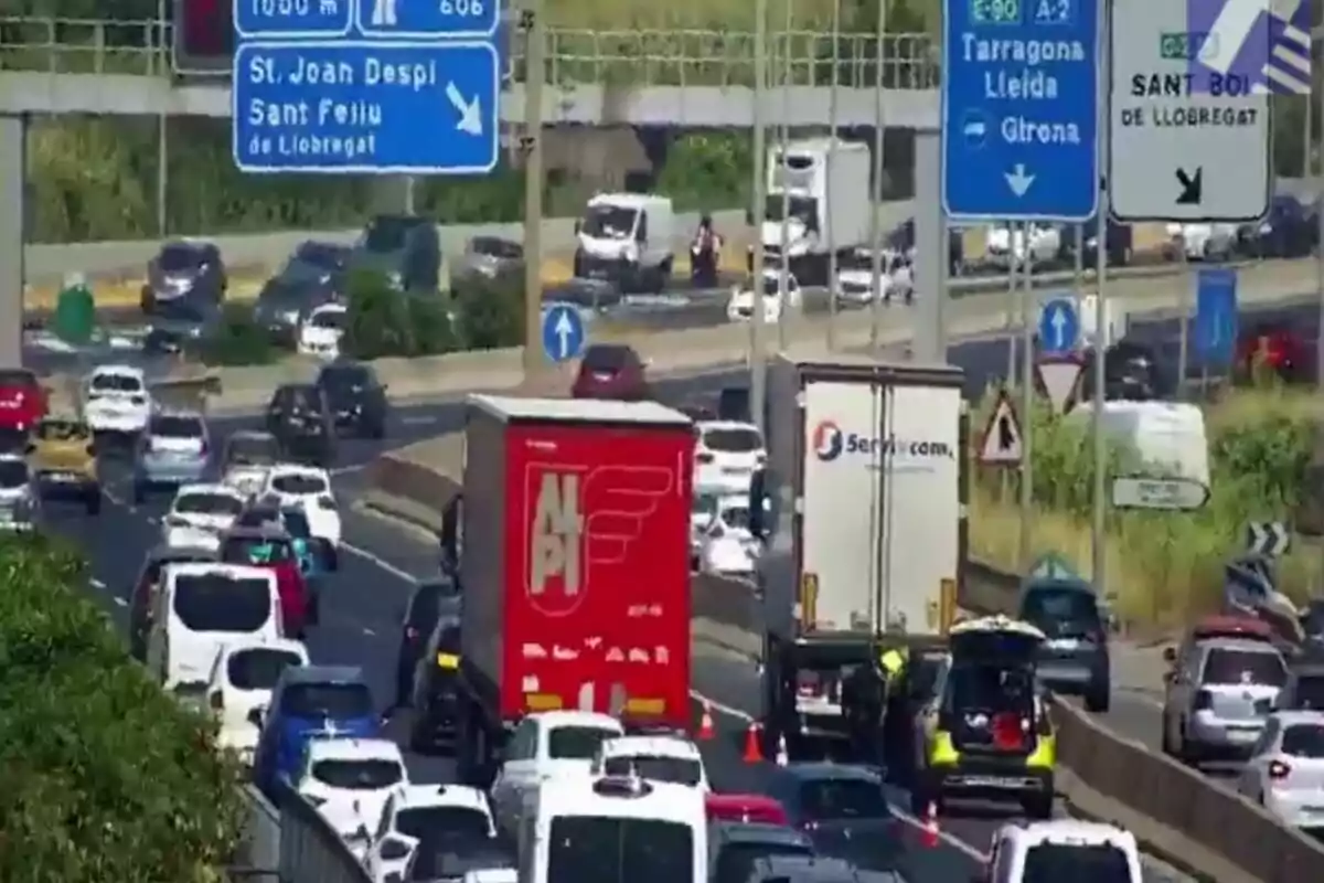 Trànsit dens en una autopista amb diversos cotxes i camions, senyals viàries blaves i blanques indicant direccions cap a Sant Boi, Tarragona, Lleida i Girona, i un vehicle d’emergència aturat al carril dret.