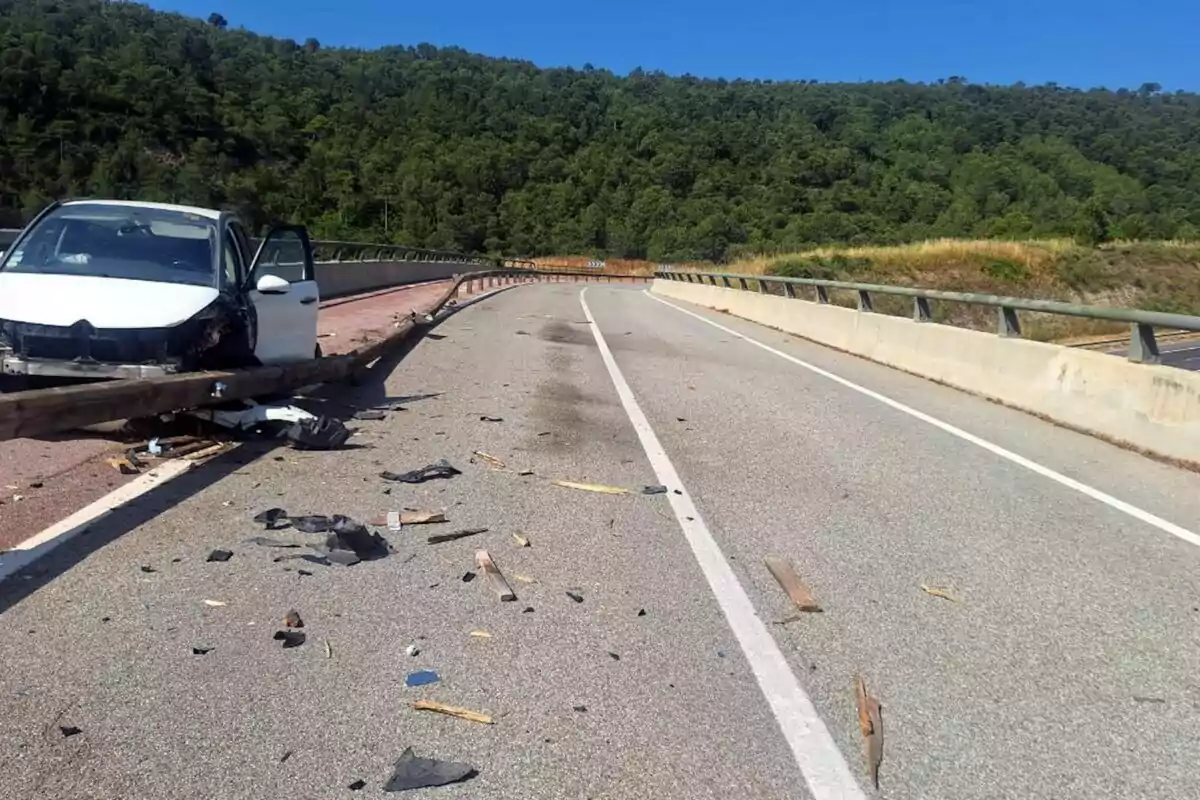 Coche blanco accidentado en un puente con escombros esparcidos sobre el asfalto y barandilla dañada, rodeado de vegetación y cielo despejado