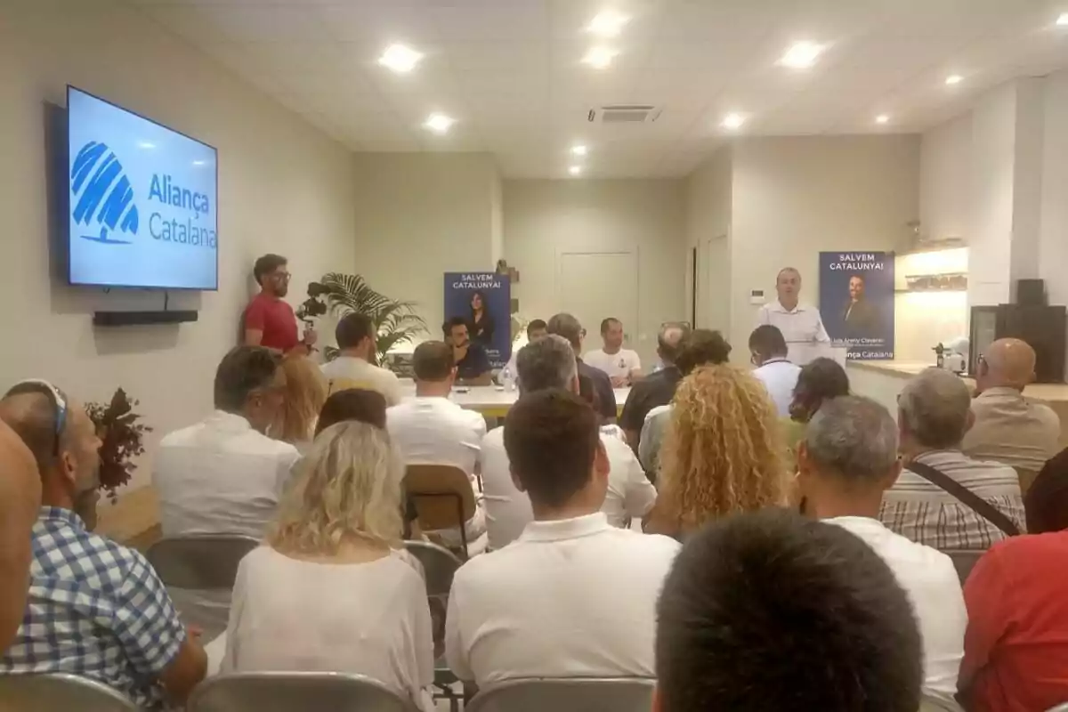 Un grup de persones assisteix a una conferència en una sala amb cartells d’Aliança Catalana i un orador dret davant del públic. Un grup de persones assisteix a una conferència en una sala amb cartells d’Aliança Catalana i un orador dret davant del públic.