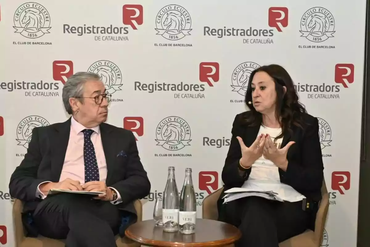 Dues persones assegudes conversen en un esdeveniment de Registradors de Catalunya amb logotips de fons i ampolles d’aigua sobre una taula petita entre elles.