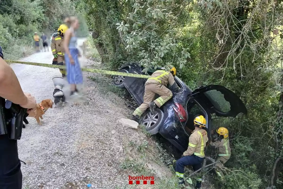 Bombers rescaten un cotxe accidentat en una rasa al costat d’un camí de terra mentre diverses persones i gossos observen l’escena.