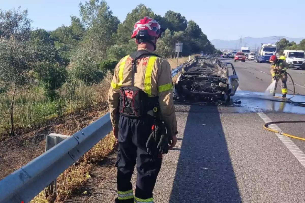 Un bomber observa un cotxe completament calcinat en una carretera mentre un altre bomber apaga les restes de l’incendi.