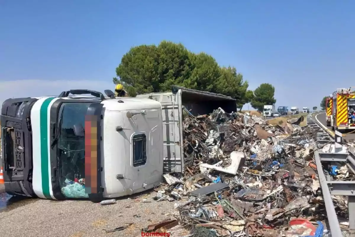 Camió bolcat a la carretera amb la seva càrrega de ferralla escampada i vehicles d’emergència atenent l’accident