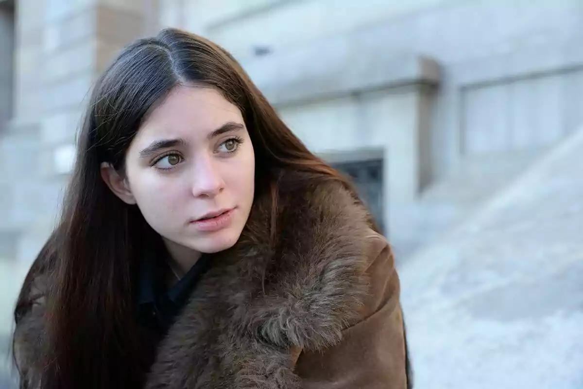 Young man with long dark hair wearing a brown faux fur coat looking to the side in an urban setting