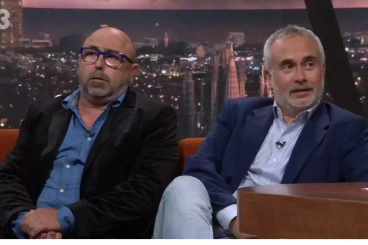 Two men sitting on a television set with a lit-up city background, both wearing jackets and jerseys, one wearing glasses and the other with a gray beard. Two men sitting on a television set with a lit-up city background, both wearing jackets and jerseys, one wearing glasses and the other with a gray beard.