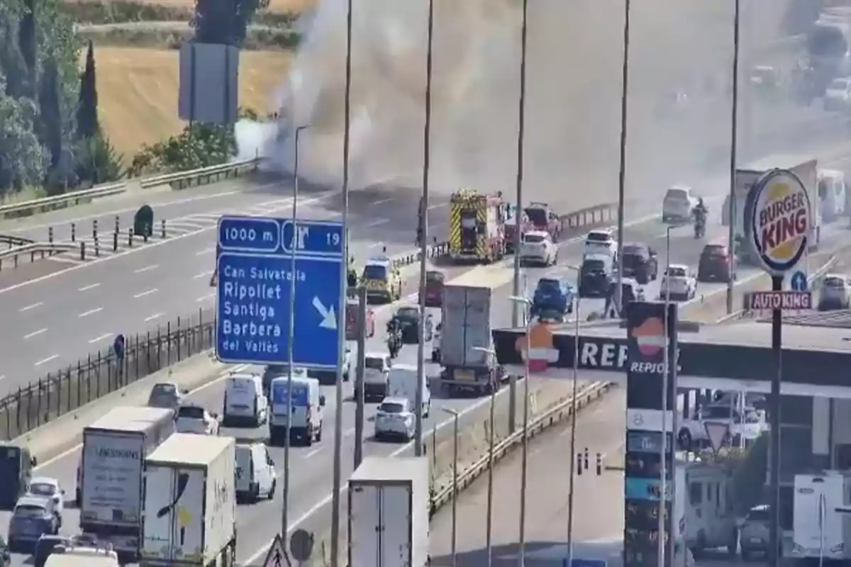 Trànsit aturat en una autopista a causa d’un incendi atès pels bombers a prop d’una benzinera i un restaurant de menjar ràpid.