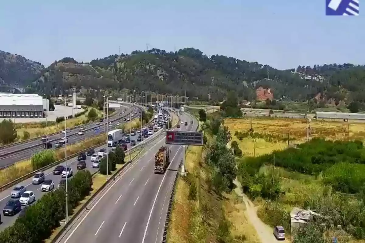 Carretera amb trànsit de vehicles envoltada de zones verdes i muntanyes al fons en un dia assolellat.