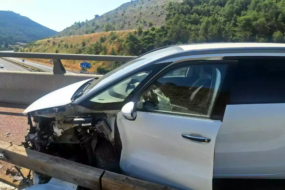 Auto blanco con la parte delantera dañada tras chocar contra una barrera de seguridad en una carretera rodeada de montañas y vegetación Auto blanco con la parte delantera dañada tras chocar contra una barrera de seguridad en una carretera rodeada de montañas y vegetación