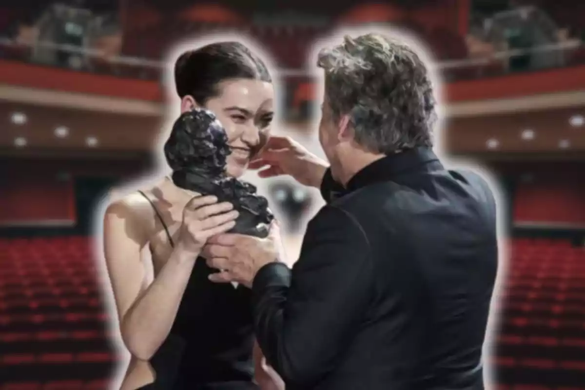 Una mujer sonríe emocionada mientras sostiene un premio en un teatro, acompañada de un hombre elegantemente vestido.
