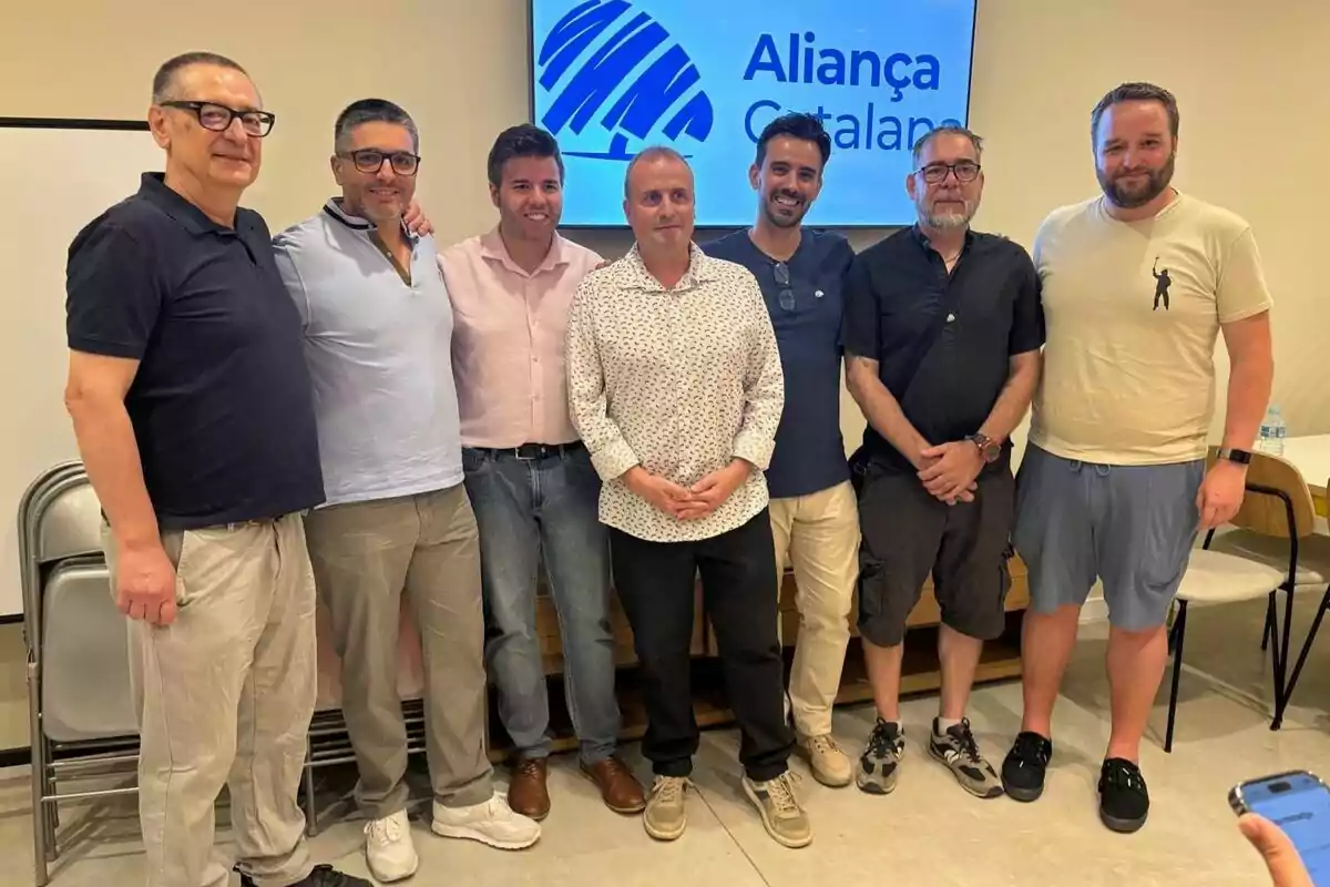 Set homes posen junts en una sala davant d’una pantalla que mostra el logotip i el nom d’Aliança Catalana.