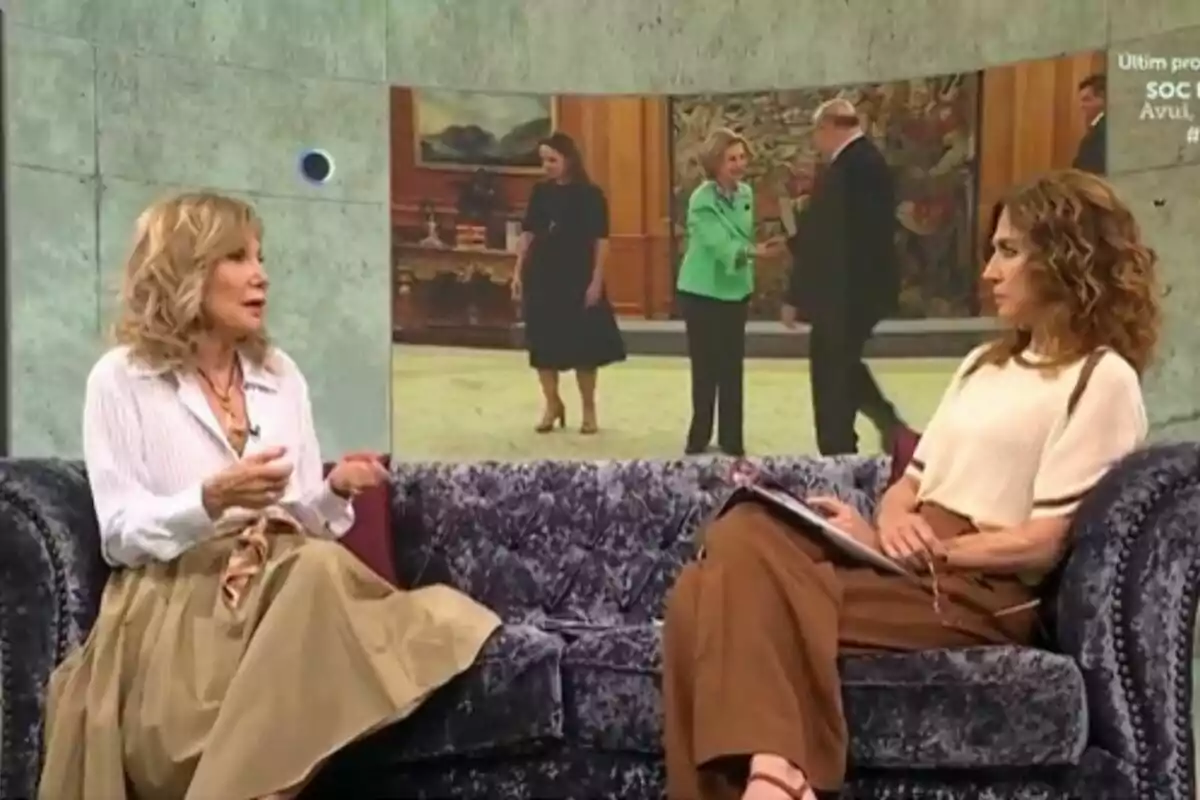 Dues dones conversen assegudes en un sofà durant un programa de televisió, amb una pantalla al fons que mostra tres persones en una sala elegant.