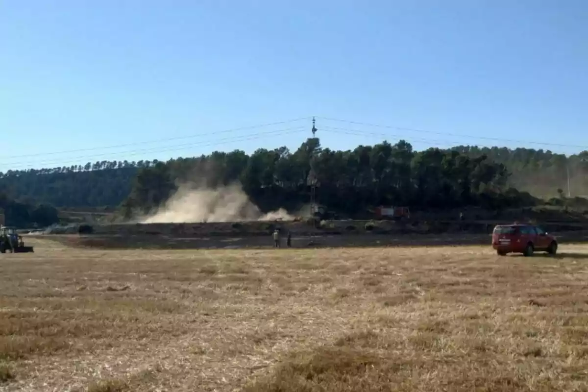 Camp sec amb fum sortint d’una zona propera a un bosc on s’observen vehicles d’emergència i persones treballant.
