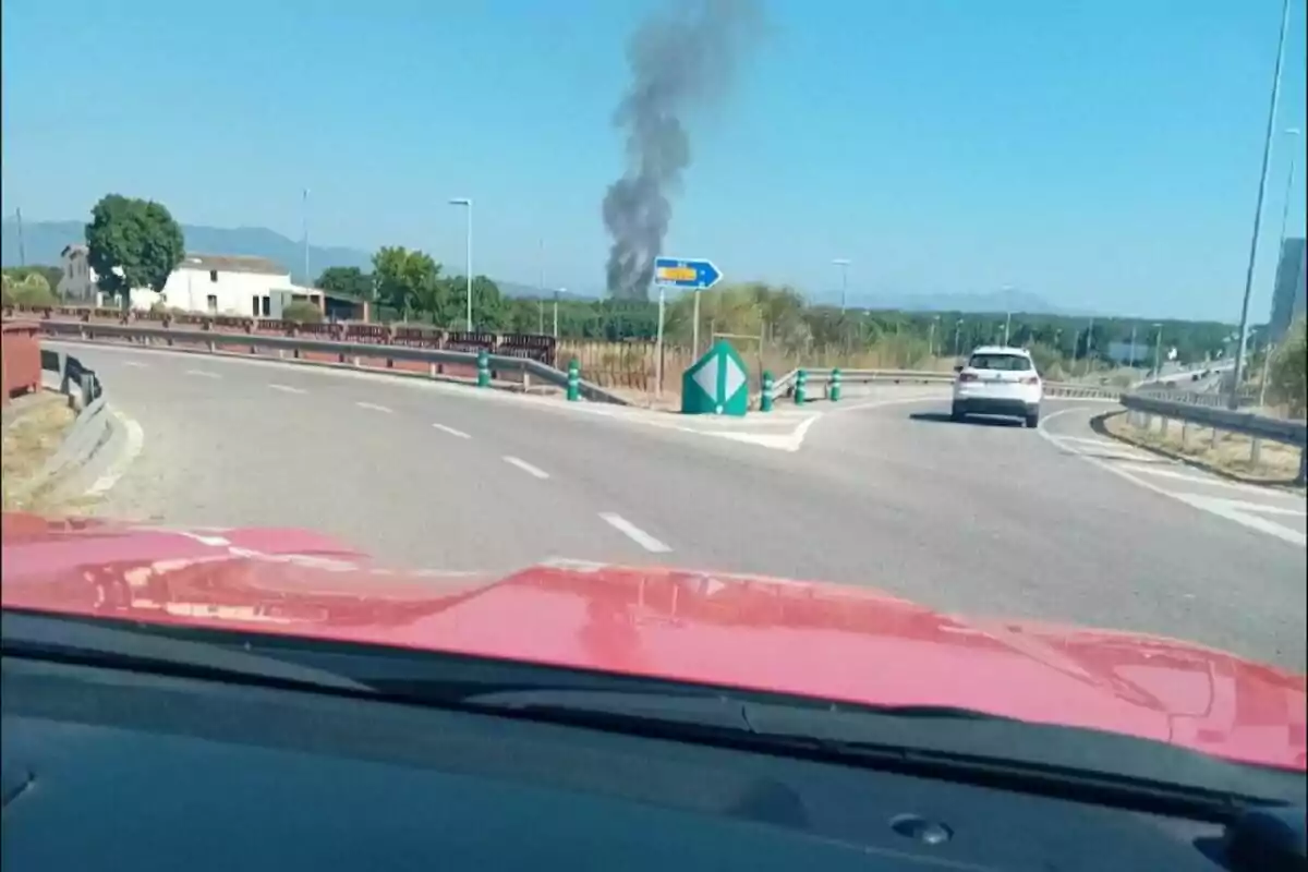 Vista des d’un cotxe en una carretera amb una columna de fum negre que s’eleva a l’horitzó i un cotxe blanc al davant