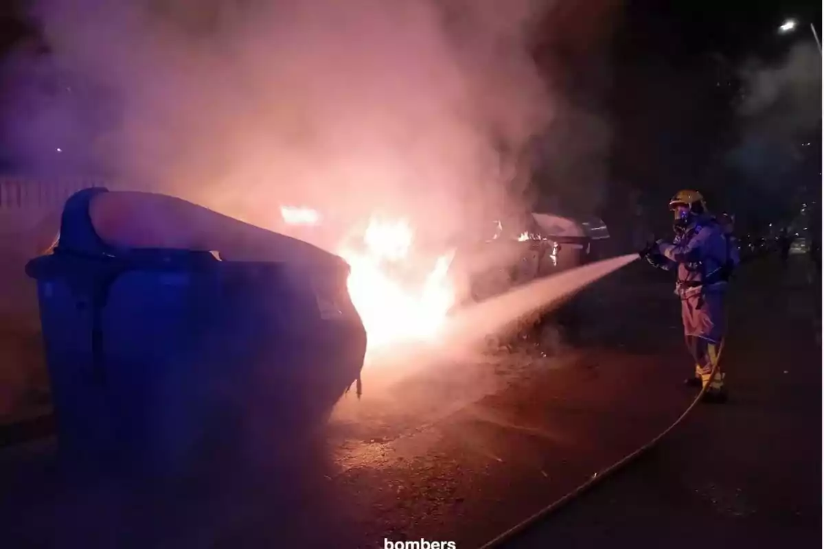 Un bomber apaga un incendi en un contenidor d’escombraries durant la nit mentre el foc i el fum s’eleven al carrer. Un bomber apaga un incendi en un contenidor d’escombraries durant la nit mentre el foc i el fum s’eleven al carrer.