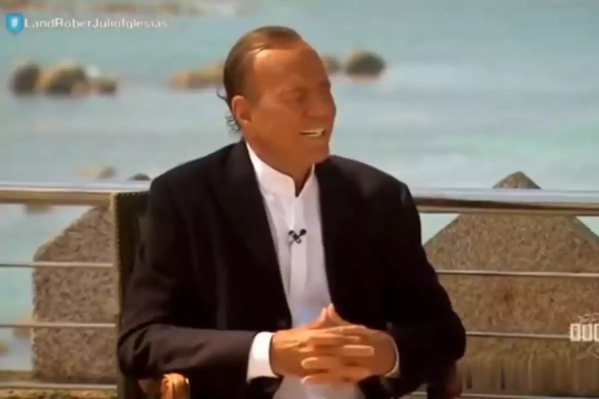 Julio Iglesias con traje oscuro y camisa blanca sentado al aire libre frente al mar sonriendo durante una entrevista. Captura de 'Land Rober'