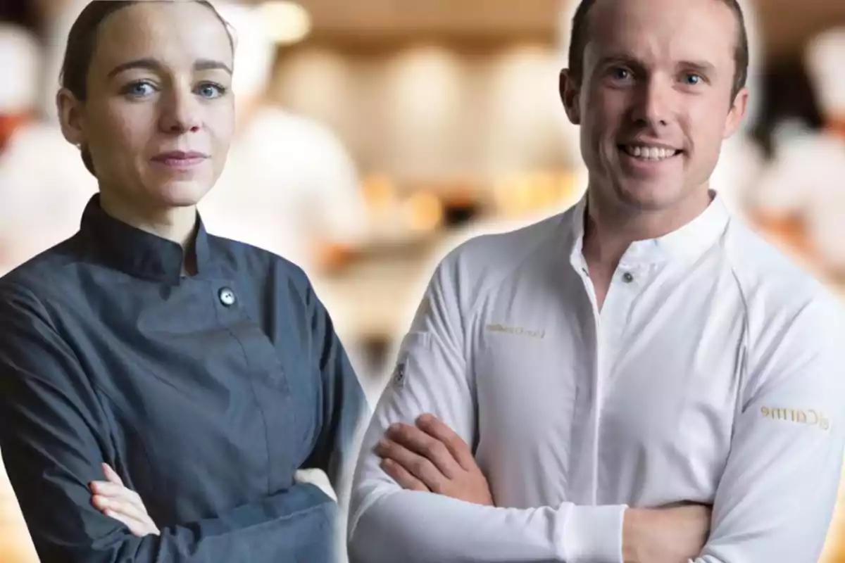 Dos chefs con uniforme de cocina posan con los brazos cruzados en un entorno de cocina profesional desenfocado.