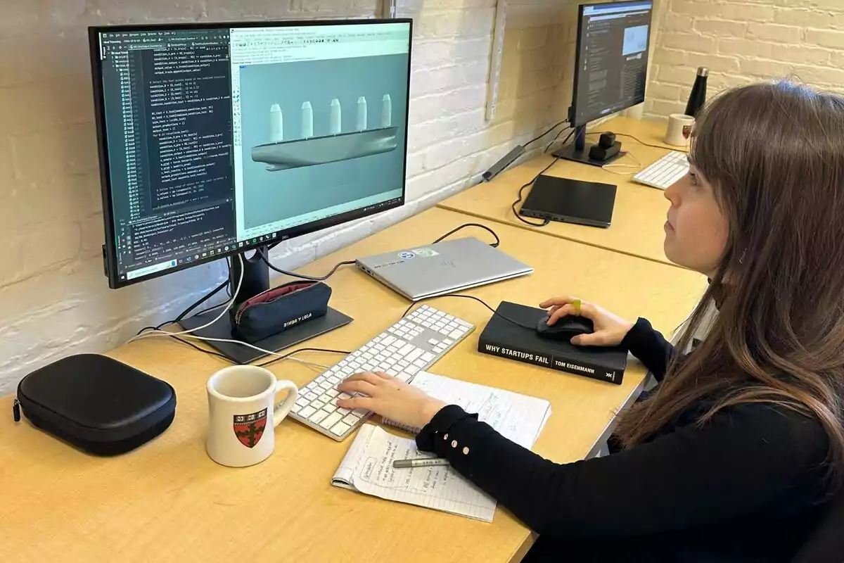 Dona treballant en un ordinador amb un monitor que mostra codi i un model 3D, envoltada d’una tassa, una llibreta, un portàtil i un llibre titulat Why Startups Fail