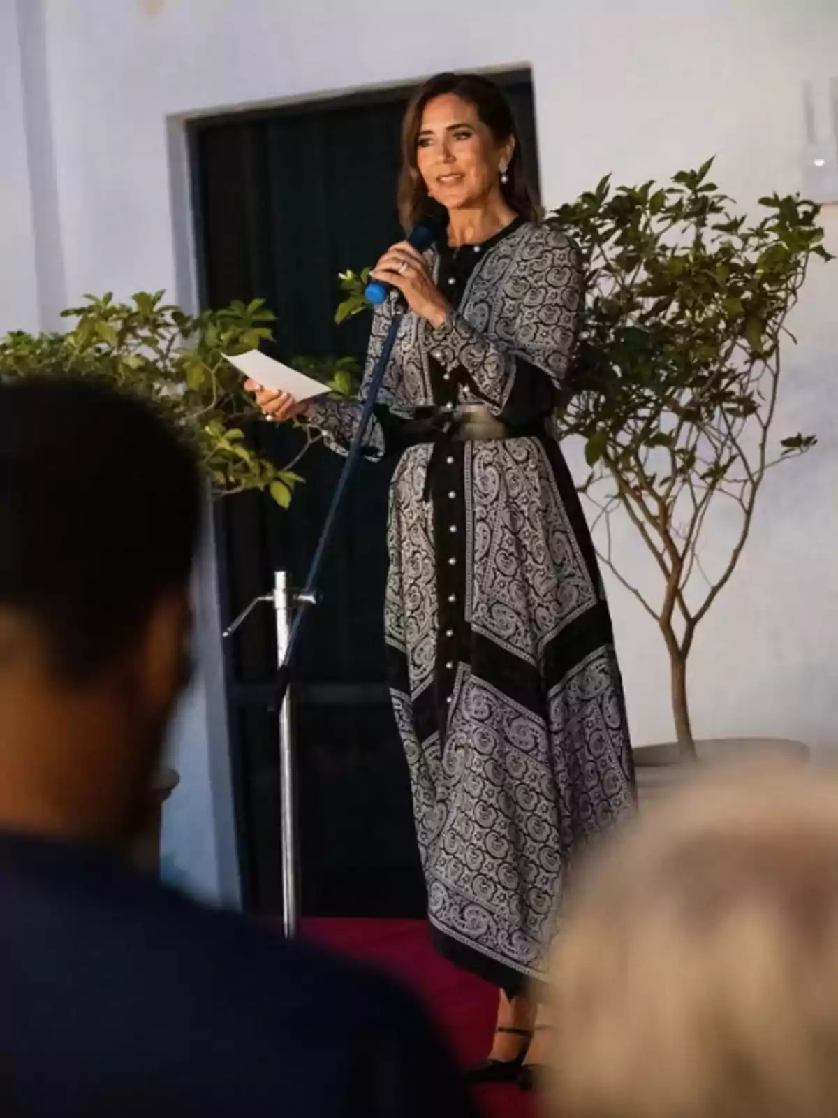 Woman standing on a platform speaking into a microphone and holding a sheet of paper while wearing a long black and white patterned dress with a black belt Woman standing on a platform speaking into a microphone and holding a sheet of paper while wearing a long black and white patterned dress with a black belt