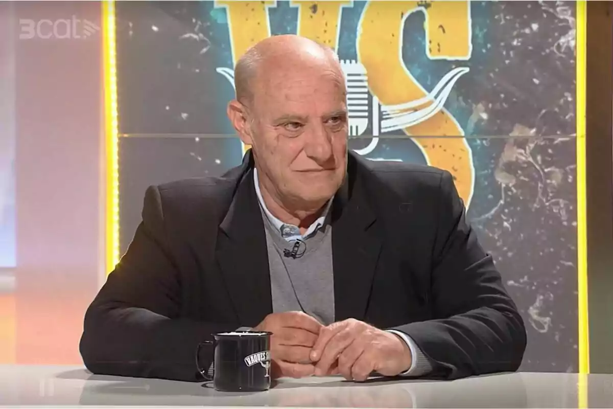 Hombre calvo de mediana edad con saco oscuro y suéter gris sentado en una mesa de un programa de televisión con una taza negra frente a él y un fondo con letras grandes y coloridas