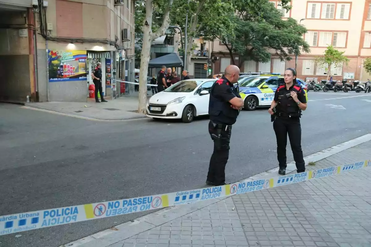 Dos agents de policia conversen en un carrer acordonat mentre altres policies treballen a prop d’un cotxe patrulla i un vehicle blanc davant d’un edifici residencial.