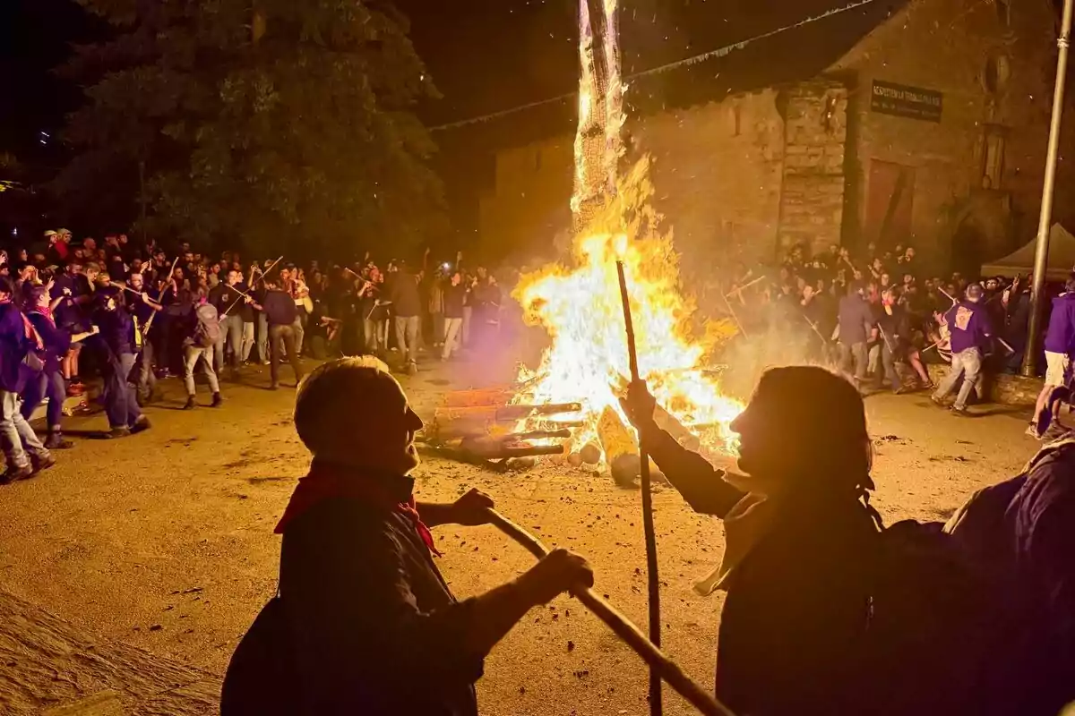 Persones celebrant al voltant d’una gran foguera en una plaça durant la nit, algunes sostenen pals i es veuen edificis antics al fons. Persones celebrant al voltant d’una gran foguera en una plaça durant la nit, algunes sostenen pals i es veuen edificis antics al fons.