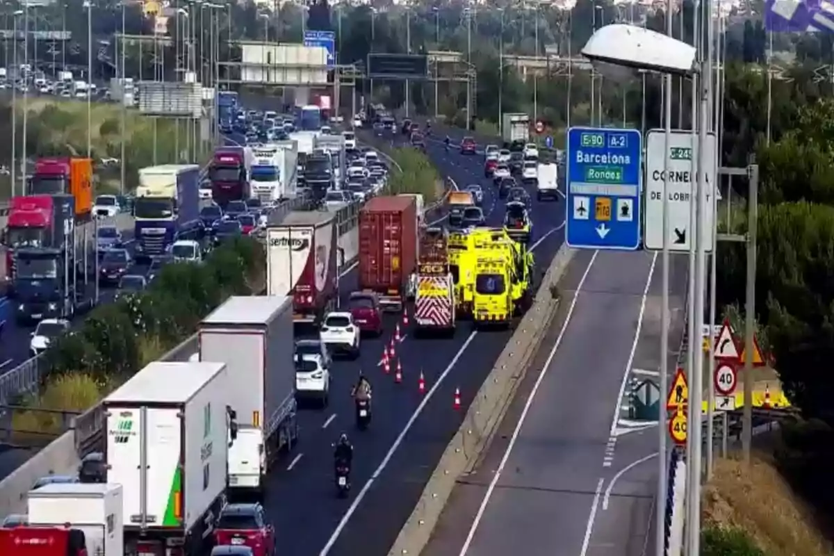 Trànsit dens en una autopista amb diversos camions i cotxes, presència d’ambulàncies i vehicles d’emergència, cons taronges delimitant un carril i senyals indicant direcció a Barcelona.