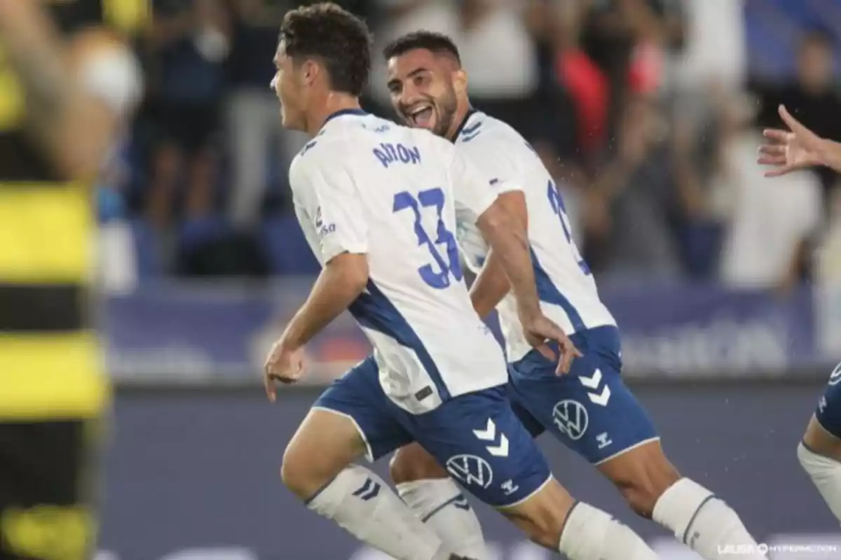 Dos futbolistes amb uniforme blanc i blau celebren un gol en un partit mentre corren somrients pel camp. Dos futbolistes amb uniforme blanc i blau celebren un gol en un partit mentre corren somrients pel camp.