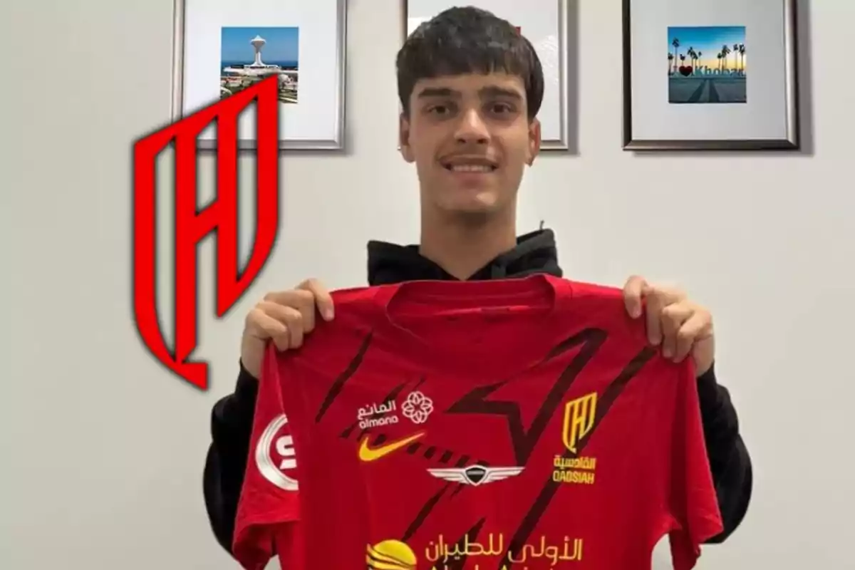 Jove somrient sosté una samarreta vermella d’un equip de futbol amb logotips i lletres en àrab, al fons hi ha quadres a la paret i un gran escut del club a l’esquerra. Jove somrient sosté una samarreta vermella d’un equip de futbol amb logotips i lletres en àrab, al fons hi ha quadres a la paret i un gran escut del club a l’esquerra.