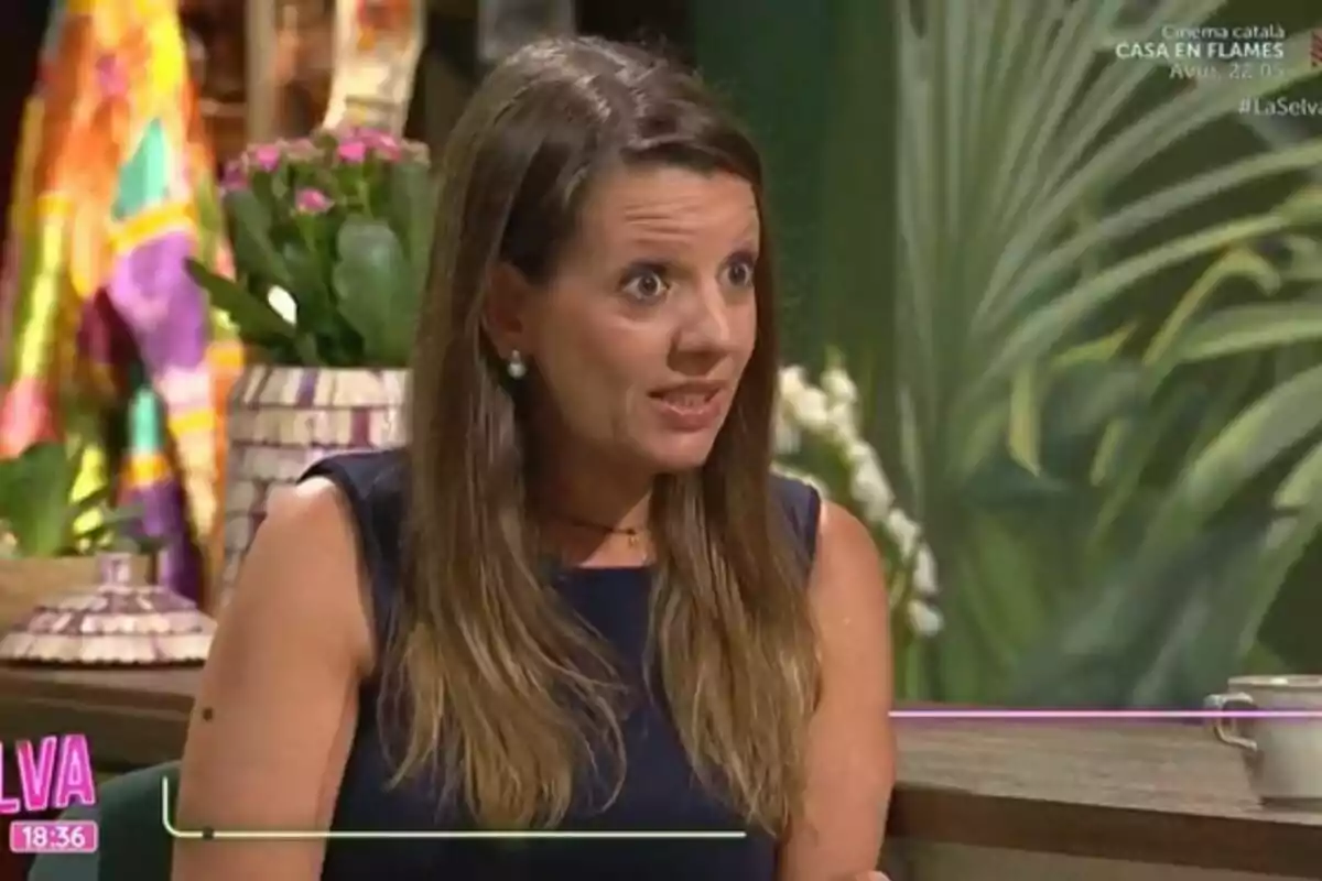 Long-haired brunette woman in a navy blue dress speaking on a set decorated with plants and vibrant colors