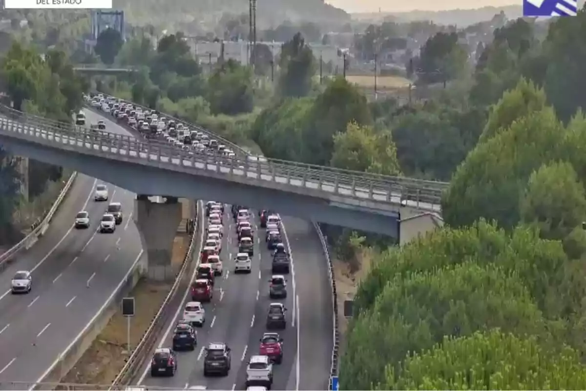 Carretera amb trànsit dens en un sentit i un pont creuant per sobre de l’autopista envoltada d’arbres