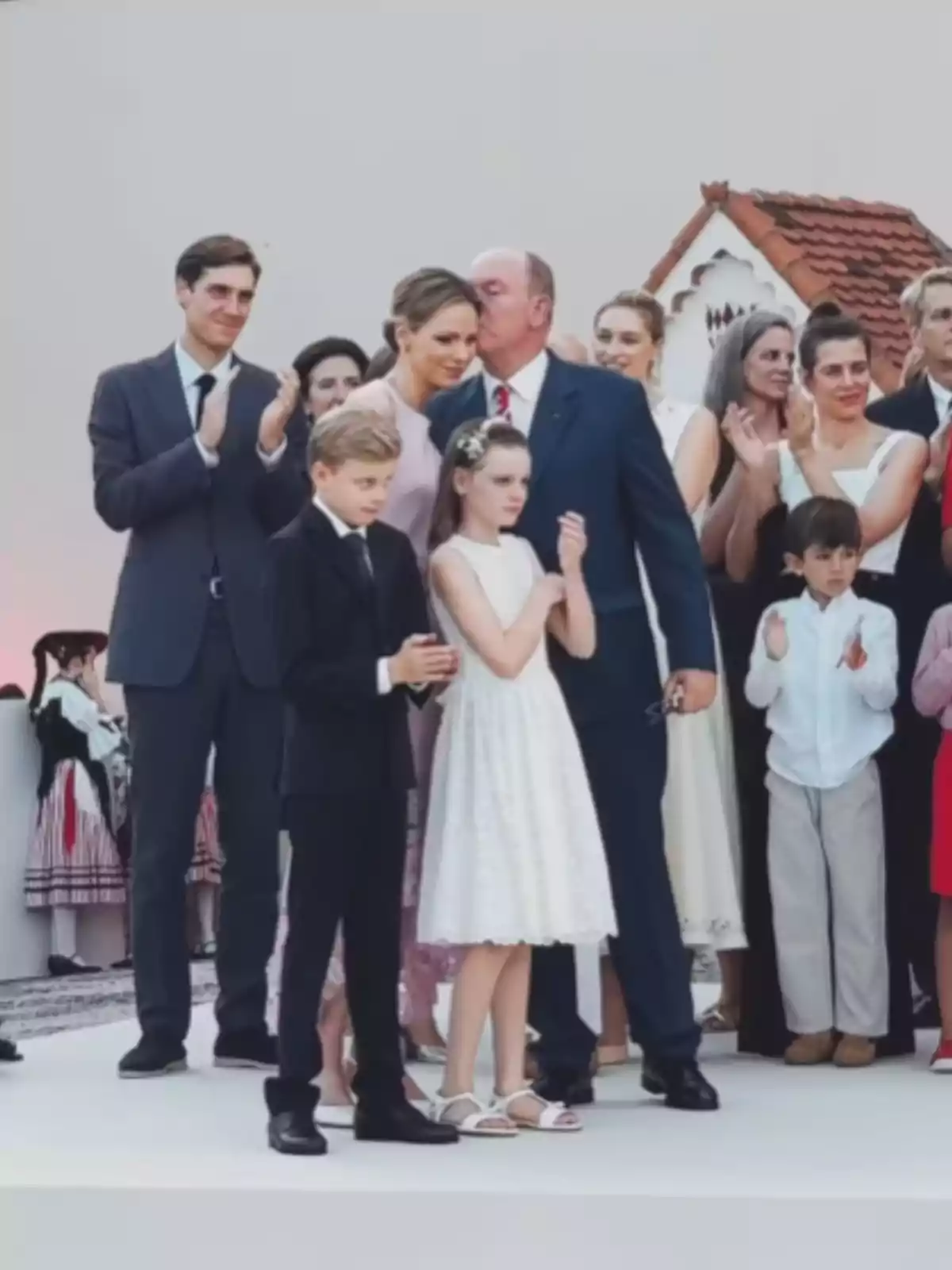 Grup de persones elegantment vestides reunides en un escenari a l’aire lliure mentre algunes aplaudeixen i un home fa un petó al cap d’una dona. Grup de persones elegantment vestides reunides en un escenari a l’aire lliure mentre algunes aplaudeixen i un home fa un petó al cap d’una dona.