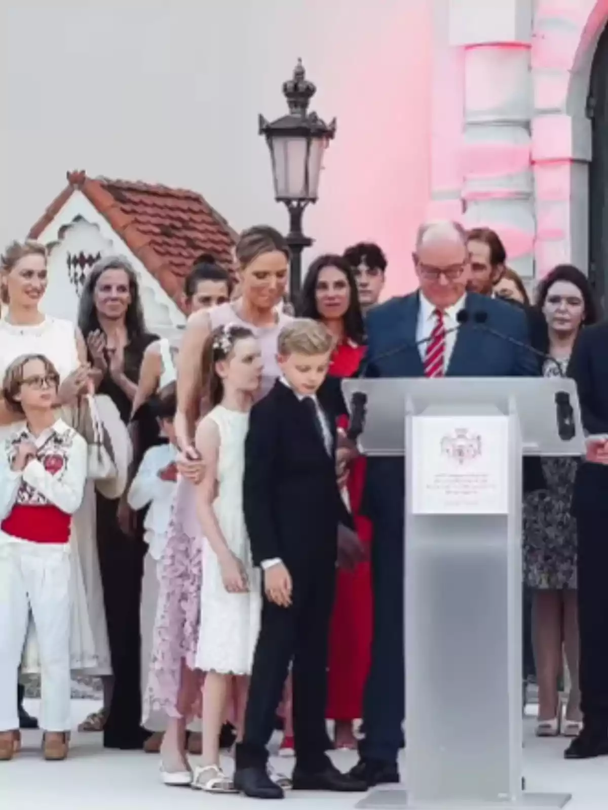 A group of elegantly dressed people gathers around a podium at an outdoor event, with an older man in a suit and tie speaking while children and adults stand with him. A group of elegantly dressed people gathers around a podium at an outdoor event, with an older man in a suit and tie speaking while children and adults stand with him.