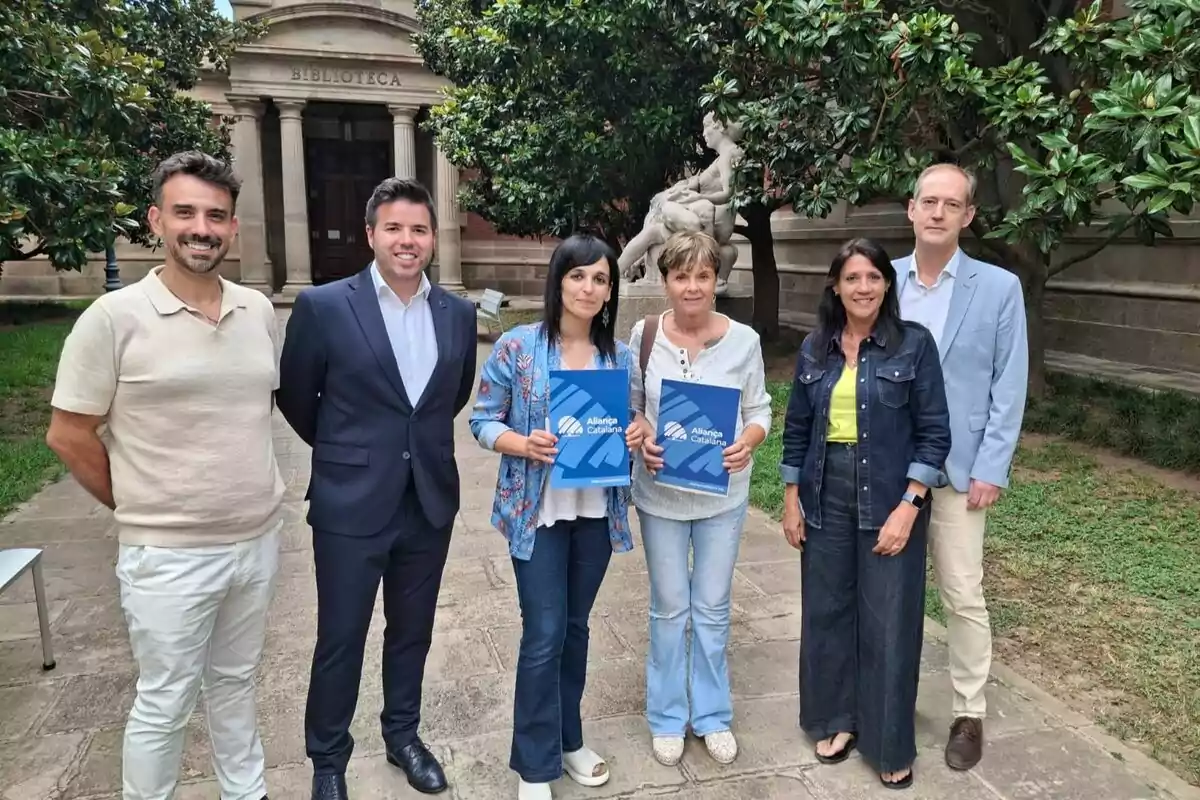Sis persones posen a l’aire lliure davant d’un edifici amb la paraula biblioteca, dues d’elles sostenen carpetes blaves amb el logotip d’Aliança Catalana, i al fons hi ha una estàtua blanca envoltada d’arbres.