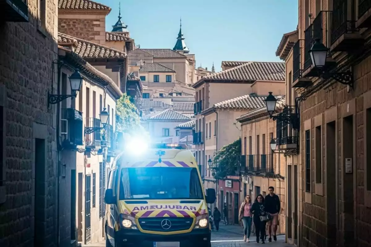Ambulància amb els llums encesos circulant per un carrer estret d’un poble amb edificis de pedra i persones caminant per la vorera Ambulància amb els llums encesos circulant per un carrer estret d’un poble amb edificis de pedra i persones caminant per la vorera