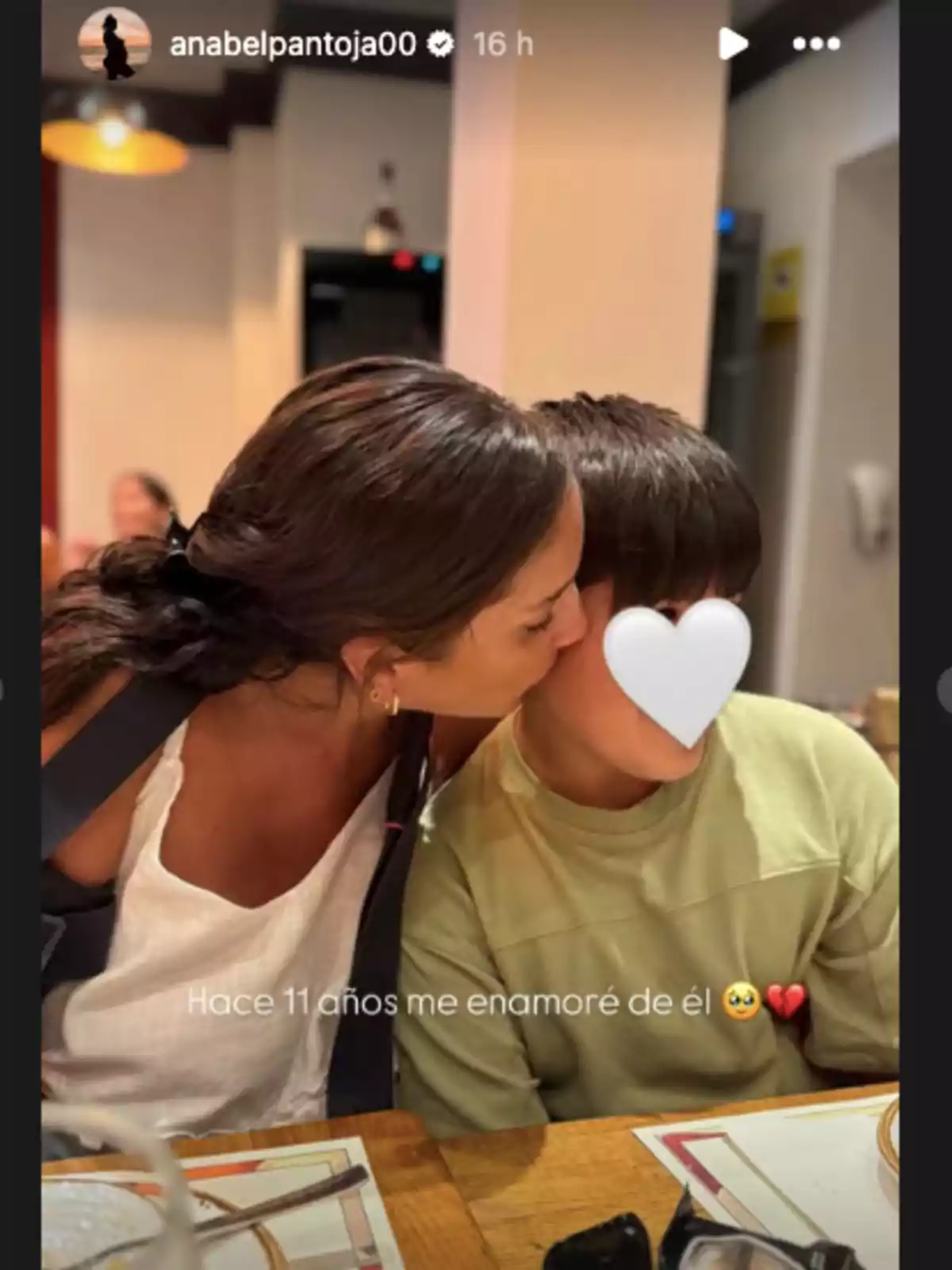 Anabel Pantoja fent un petó a la galta d’un nen amb la cara tapada amb un emoji de cor blanc mentre estan asseguts en una taula de restaurant Anabel Pantoja fent un petó a la galta d’un nen amb la cara tapada amb un emoji de cor blanc mentre estan asseguts en una taula de restaurant