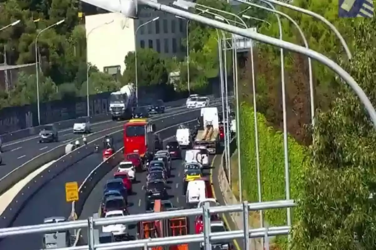 Tráfico denso en una autopista con varios autos, camiones y un autobús rojo avanzando lentamente junto a una pared cubierta de vegetación.