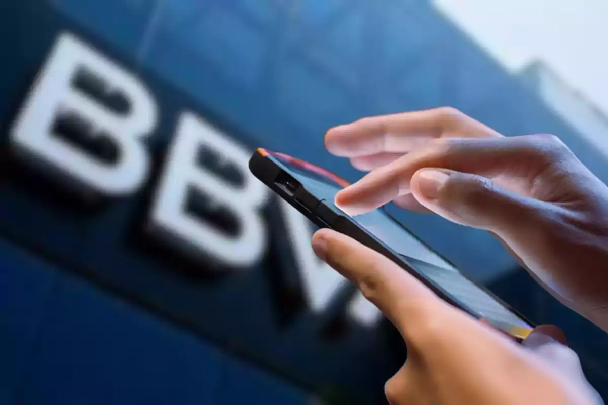 Persona utilitzant un telèfon mòbil davant d’un edifici amb el logotip de BBVA desenfocat.