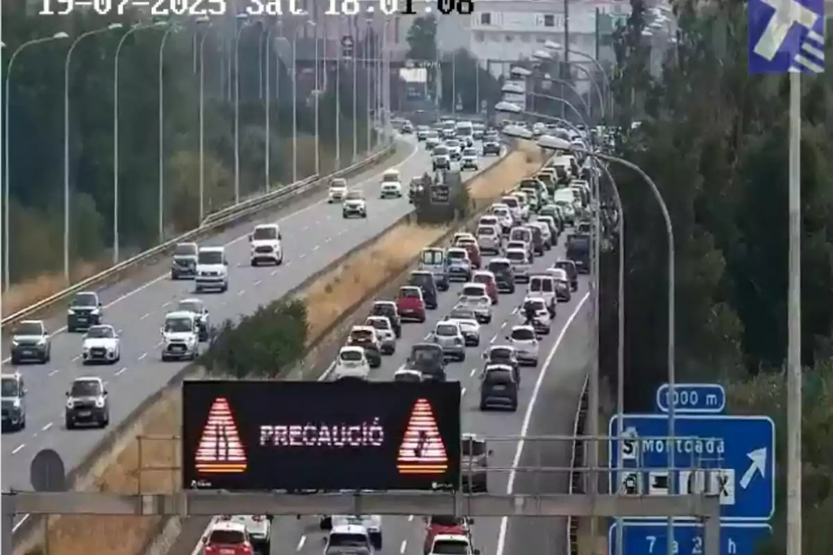 Tráfico denso en una autopista con una señal luminosa que indica precaución y un cartel azul señalando la salida hacia Montcada