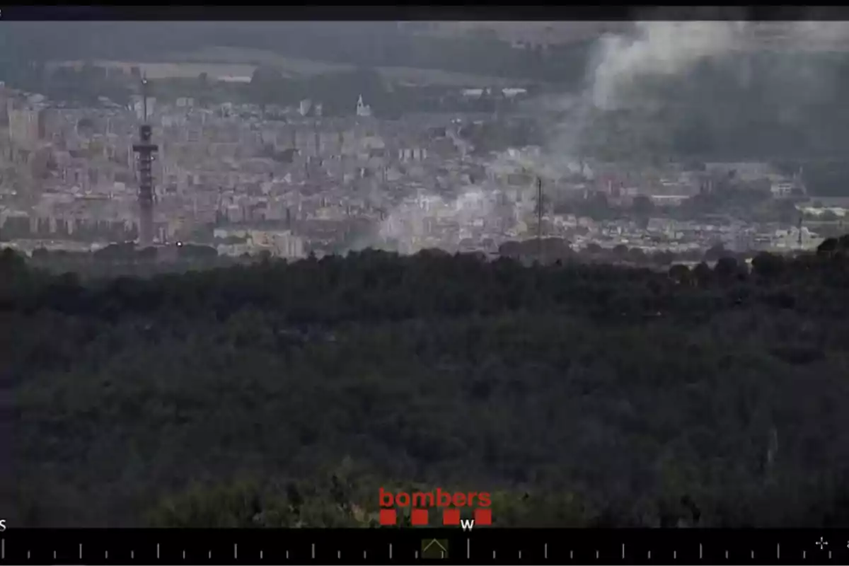 Vista panoràmica d’una ciutat amb una columna de fum que s’eleva a la zona urbana i una àrea boscosa en primer terme