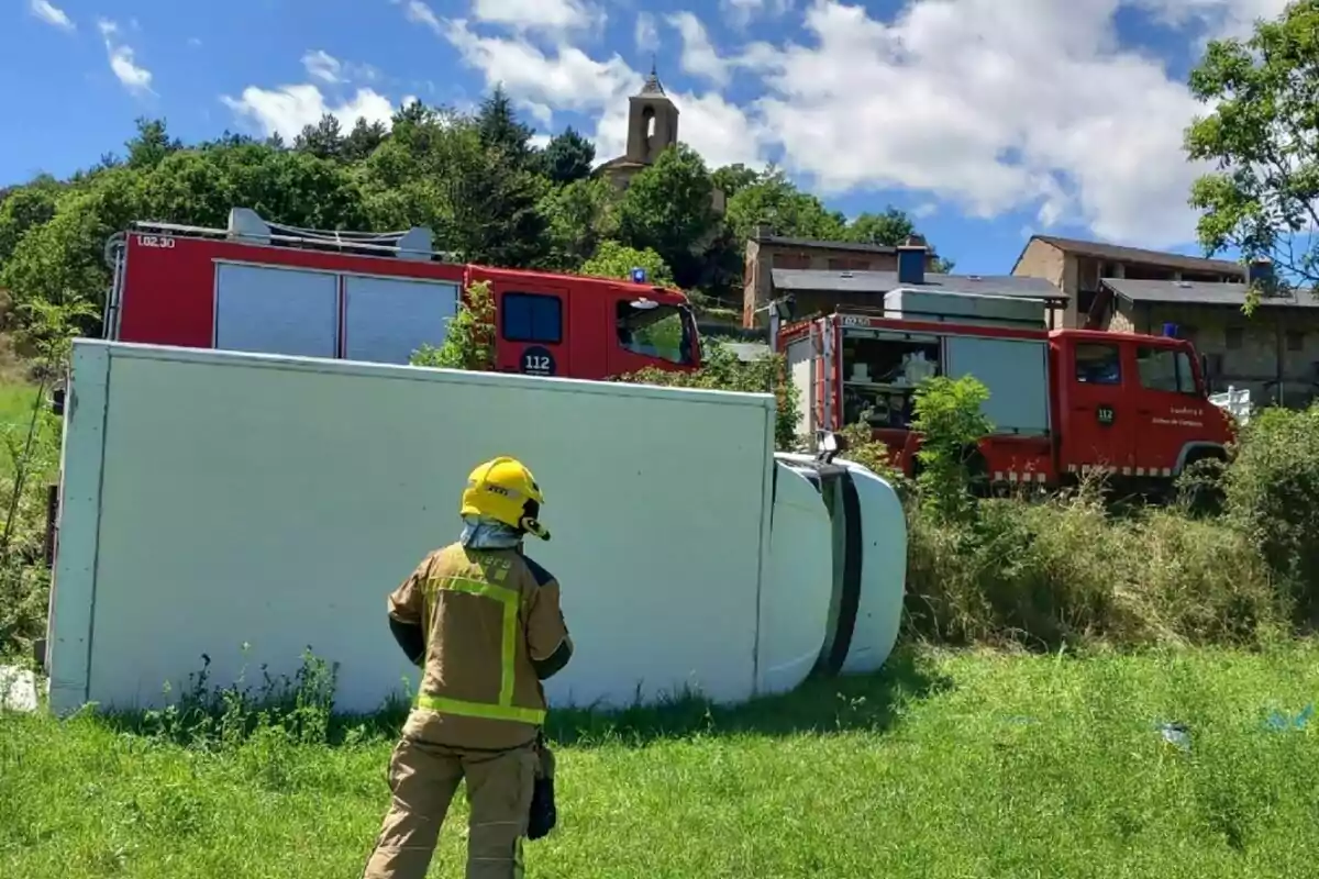 Un bombero observa un camión volcado en un campo mientras dos camiones de bomberos están estacionados cerca de unas casas rurales