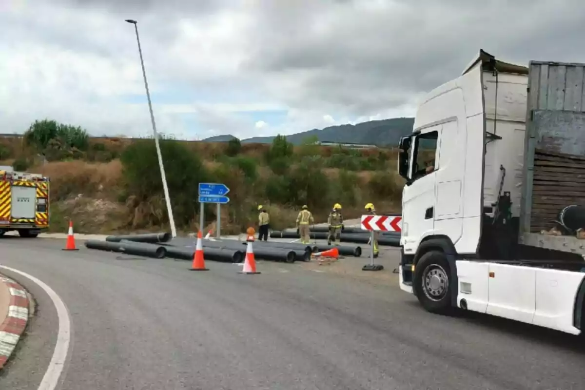 Un camió aturat en una corba amb tubs caiguts a la carretera i diversos treballadors amb casc i armilla a prop d’un camió de bombers