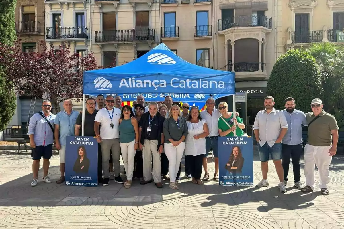 Un grup de persones posant davant d’una carpa blava d’Aliança Catalana en una plaça, amb cartells de campanya i edificis al fons