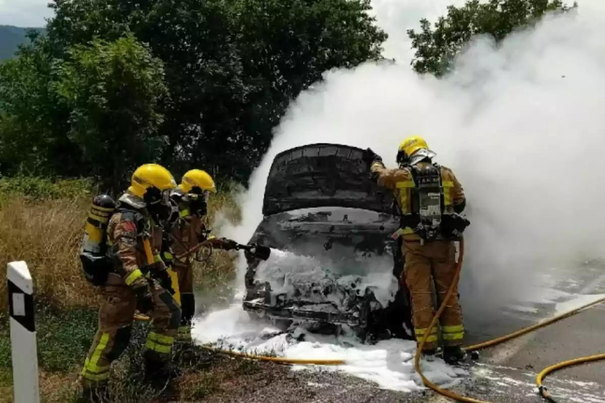 Tres bomberos apagan el incendio de un coche en una carretera usando espuma mientras el vehículo está cubierto de humo blanco