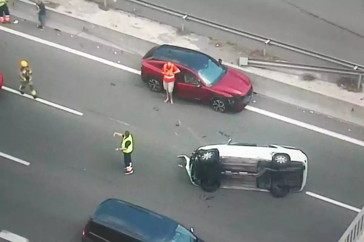 Accident viari en una carretera amb un cotxe bolcat de costat, un vehicle vermell aturat i diverses persones, inclòs personal d’emergència, atenent la situació.