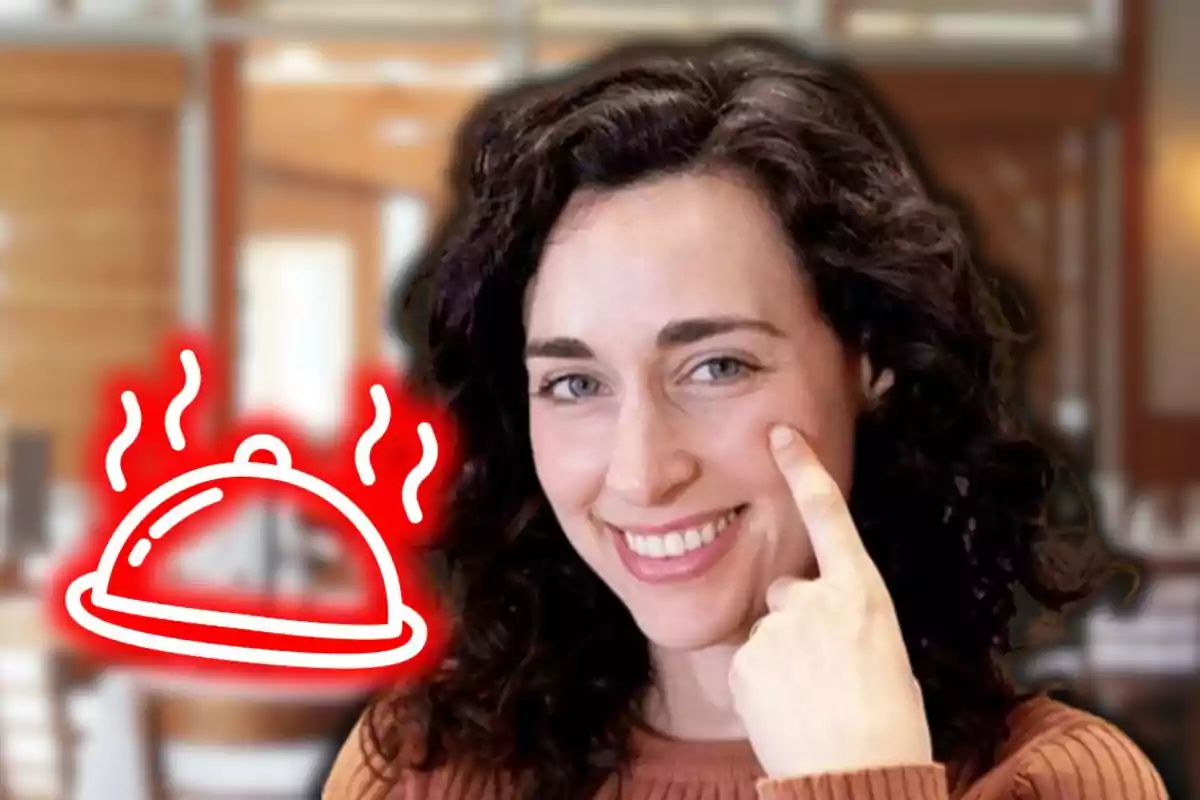 Dona de cabells arrissats somrient i assenyalant la galta amb un dibuix d’una safata de menjar fumejant al seu costat en un ambient de restaurant desenfocat