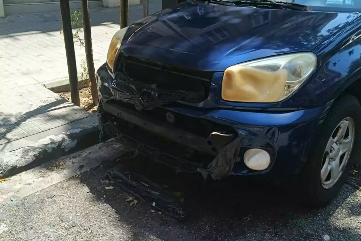Cotxe blau amb la part davantera danyada i el para-xocs desprès estacionat al costat de la vorera Cotxe blau amb la part davantera danyada i el para-xocs desprès estacionat al costat de la vorera