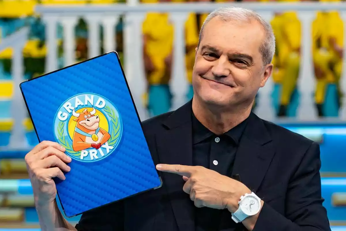 Ramón García somrient amb cabells canosos sosté i assenyala una carpeta blava amb el logotip de Grand Prix que té una vaca caricaturitzada. Ramón García somrient amb cabells canosos sosté i assenyala una carpeta blava amb el logotip de Grand Prix que té una vaca caricaturitzada.