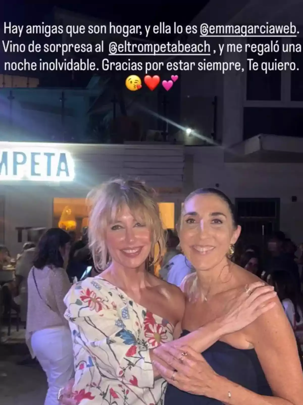 Emma García i Paz Padilla somriuen abraçades en una terrassa nocturna amb ambient festiu, una d’elles porta un vestit amb estampat floral i l’altra un vestit fosc, al fons es veu un rètol il·luminat i diverses persones gaudint de la nit. Emma García i Paz Padilla somriuen abraçades en una terrassa nocturna amb ambient festiu, una d’elles porta un vestit amb estampat floral i l’altra un vestit fosc, al fons es veu un rètol il·luminat i diverses persones gaudint de la nit.