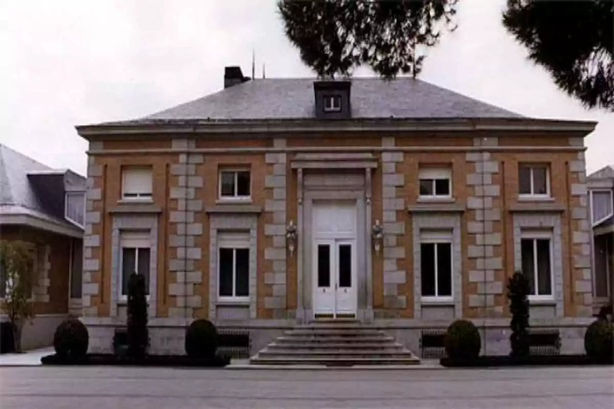 Façana d’un edifici de maó amb detalls de pedra, porta principal blanca i escales al davant envoltat de matolls i arbres. Façana d’un edifici de maó amb detalls de pedra, porta principal blanca i escales al davant envoltat de matolls i arbres.